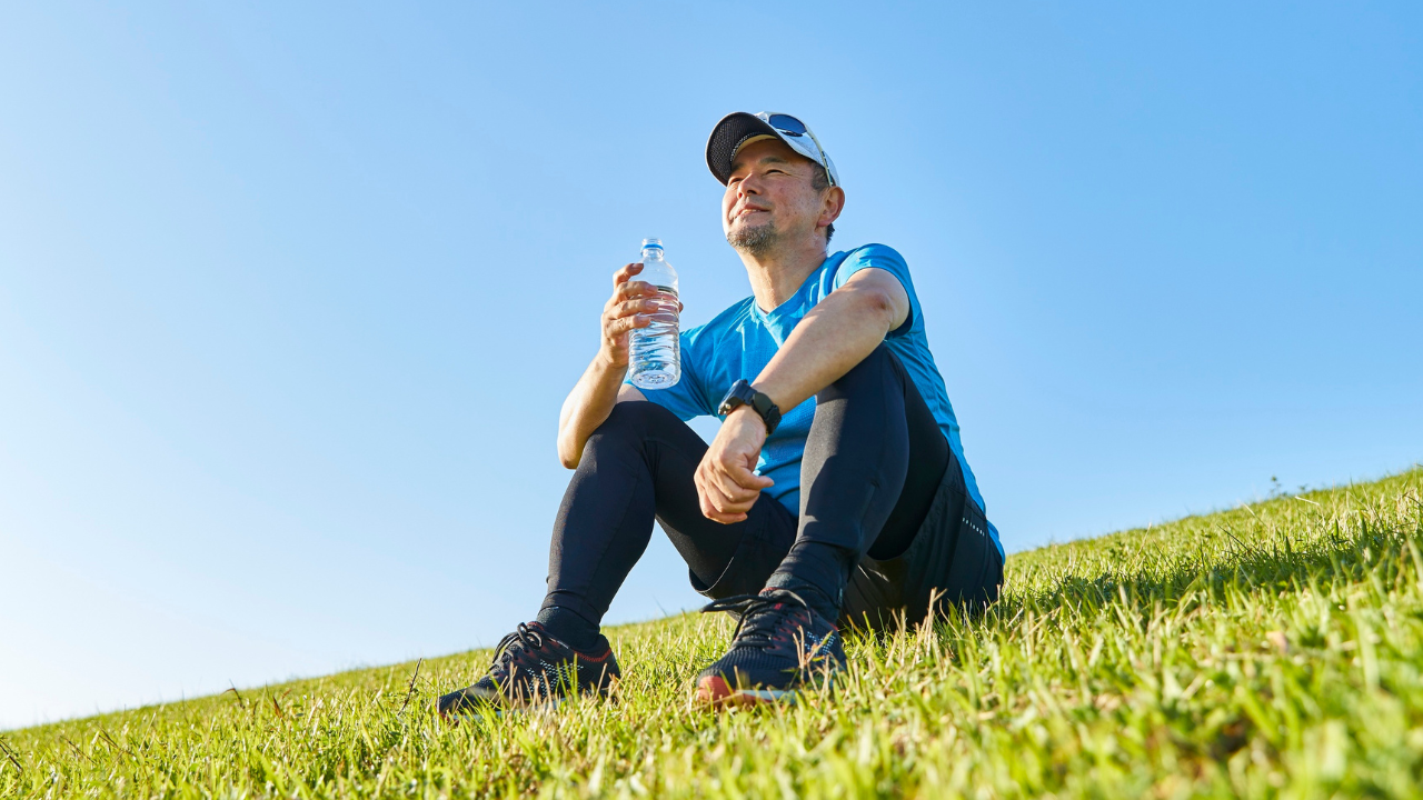 Runner sitting on a grassy hillside after a run, holding a water bottle and looking thoughtfully into the distance beneath a clear blue sky.