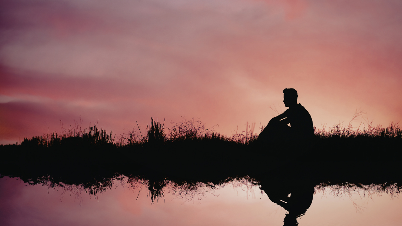 Silhouette of a person sitting quietly near water at sunset, reflected against a soft pink and purple sky.