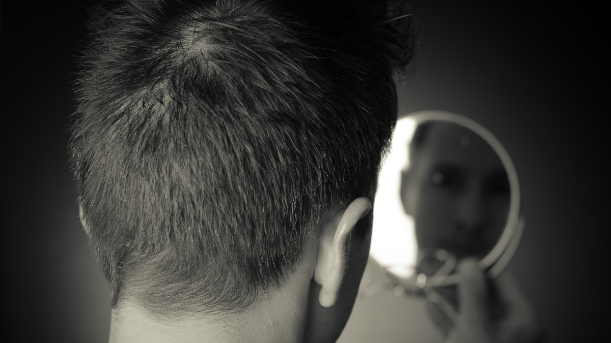 Black-and-white photograph of a person seen from behind looking into a small mirror, with their reflection softly out of focus, suggesting quiet self-reflection and questions of identity.